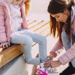 From above of young woman putting roller skates on little daughter sitting on bench during active leisure while spending time together in cold season