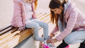 From above of young woman putting roller skates on little daughter sitting on bench during active leisure while spending time together in cold season
